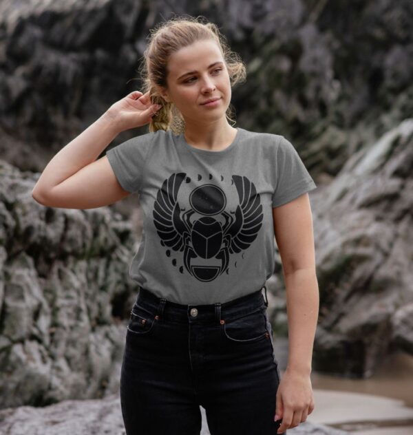 Woman by rocks on beach wearing the Winged Scarab Organic Cotton Crew Neck Tee in Mauve Color