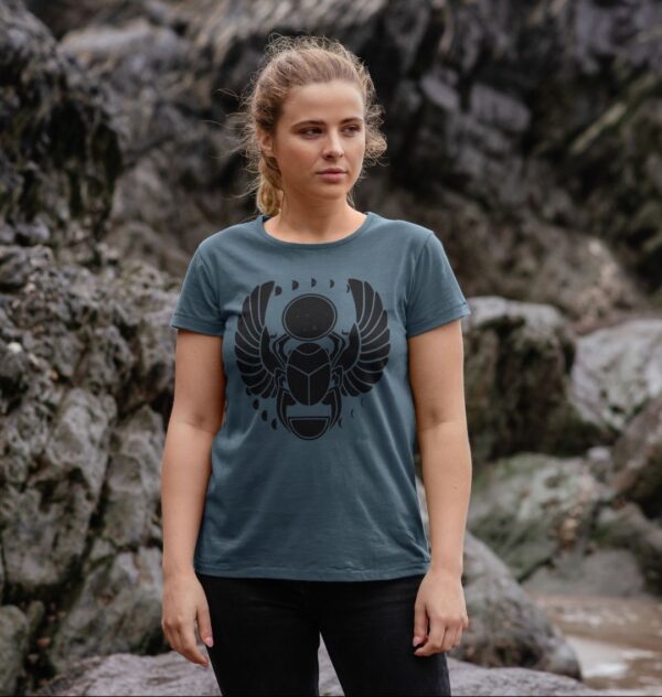 Woman by rocks on beach wearing the Winged Scarab Organic Cotton Crew Neck Tee in Mauve Color