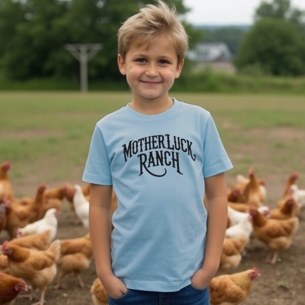 Boy wearing a blue organic cotton t-shirt that says "Mother Luck Ranch" in an old west font. He is standing in front of chickens.